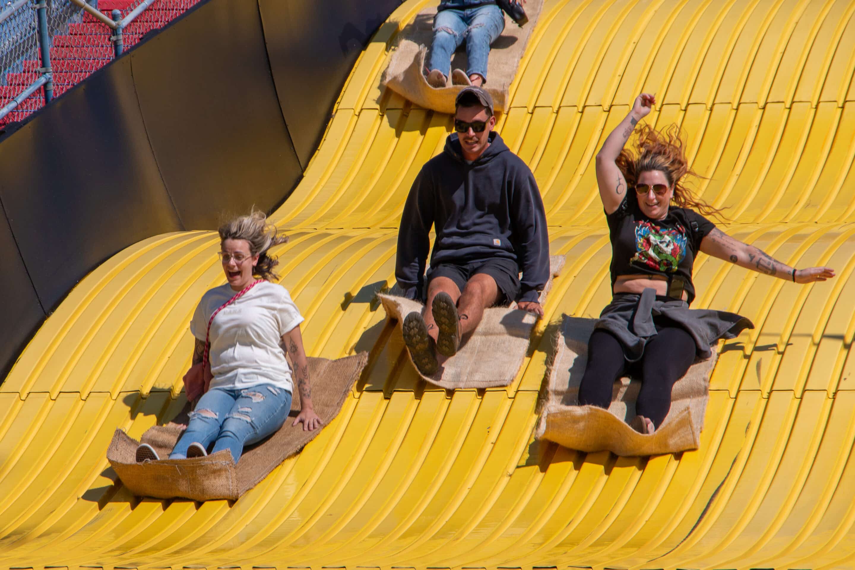 Three people sitting on potato sacks sliding down a giant yellow slide at the Big E