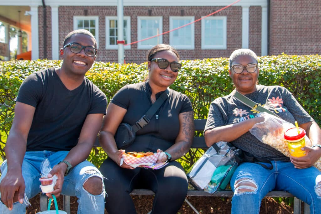 Three people with glasses holding food while sitting on a bench at the Big E